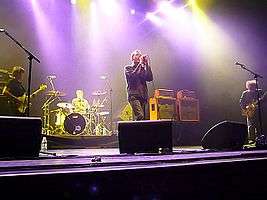 A man wearing a black jacket sings on a stage illuminated with yellow and purple lights; a bassist, a guitarist and a drummer perform along with him.