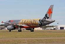 Left side on view of an aeroplane. It is basically silver in colour with standard logo of the airline. Added to the side of the plane is a large print of 'Powderfinger' in black-bordered yellow to orange-brown colouring. It spans the distance from where the wings join to just before the rear doorway. To the right and just below the band's name, in smaller lettering is the word 'Sunsets', coloured mostly in brown. Either side of the runway are grassed areas, beyond the jet are buildings – mostly obscured.