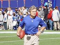 Candid photograph of McNally wearing a blue t-shirt bearing a New York Giants logo standing on a football field and holding a football in his right hand with his left hand on his hip
