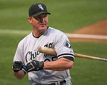A man in a grey baseball uniform with "Chicago" on his chest in black with a black cap, black batting gloves, and a baseball bat under his arm.