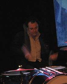 A 45-year-old man is shown at his drum kit. Both drum sticks are blurred in motion. Part of the drum kit is out of view. The man is dressed in a dark jacket with a light shirt, his eyes are focussed down. Behind him is a mostly dark background, past his left shoulder is a blue and white picture.