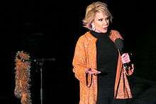 A woman with short blond hair wearing black except for a sparkly orange jacket with earrings. She is holding a microphone, and next to her is a microphone stand with an orange boa hanging from it.