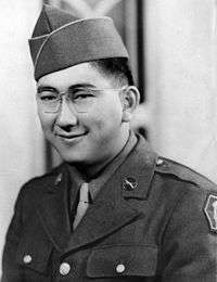 Head and shoulders of a smiling young man with dimples and round wire-framed glasses wearing a garrison cap and a military jacket over a shirt and tie.