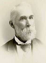 A white-haired man in his early sixties with a white mustache and beard. He is wearing a black coat, white shirt, and black tie