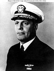 Head and shoulders of a middle aged white man wearing a white peaked cap with a black visor and a dark jacket over a white shirt and dark tie. On his left breast is a winged pin and a single ribbon bar.