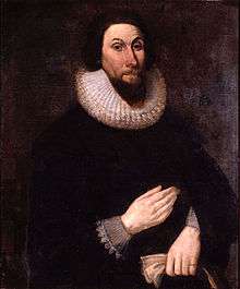 A painting of a man with a stern expression on his face, wearing very dark clothing so that his pale hands show boldly.  His hands are placed in front of him, separately, one above the other.