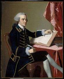 Full-length portrait of a young man seated at a table - he wears a finely tailored dark suit, knee breeches with white stockings, and a wig in the style of an English gentleman. He holds a quill pen in his right hand, and is turning the pages of a large book with the other hand.