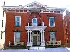 A brick house with white ornate wooden trim seen from the front. Its roof has a small pointed section in the middle, and the front door is boarded over. There is snow on the ground in front and the steps.