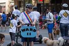 A man in a T-shirt and sunglasses, with a rainbow sash and bicycle helmet, rides a Segway adored with a sign reading "Ward 11".