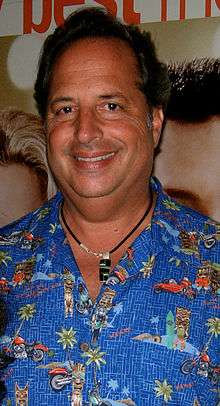 A black-haired man with a blue based shirt and a necklace looks straight at the camera smiling.