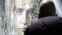 The face of a man appears in a crystalline wall, while a bald man with his back at the camera looks at him.