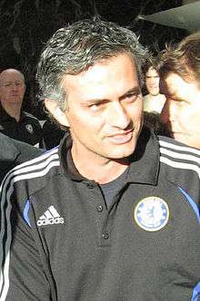 The head and upper torso of a man with black, greying hair. He is wearing a black polo shirt with the blue Chelsea F.C. crest on the left breast and a white logo of the Adidas sponsor on the right breast. Three white stripes are visible on the shoulder.