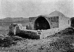 A black-and-white photograph showing a low stone wall enclosing a courtyard in front of a low building with an entry through a pointed arch with a small dome behind