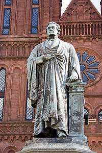 An aged bronze statue of a man wearing a robe. His left hand rests on a book which sits on a pedestal and the statue is in front of a red brick building reminiscent of a Romanesque Cathedral