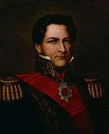 Half-length painted portrait of a man with curly hair, long sideburns and blue eyes who wears a heavily embroidered military tunic with high collar, gold braid epaulettes and a red sash of office