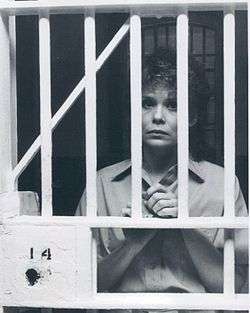 A women with short hair, wearing shirt, while holding the bars of a fictional prison.