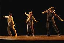 Three men in hats and beige or brown outfits dance on a dark stage