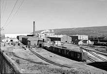 Black-and-white photo showing tracks and box cars waiting outside rows of rectangular shop buildings, one with a smoke stack.