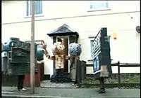 A person dressed in a cardboard costume with a large cardboard head and arms exits the central door of a two storey house which is painted cream. A short path leads directly from the door through a small garden to the pavement outside. To the left of the path a man dressed as a camcorder stands on the pavement behind a lamppost, facing the doorway. At the very end of the path, to the right, a man dressed as a microphone stands sideways, looking at the doorway. On the pavement to the right of the path a person dressed as a clapperboard stands looking down the pavement towards the person dressed as a camcorder.
