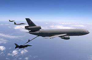 Large three-engined aircraft refueling a jet fighter while two more of the latter fly in the distance.