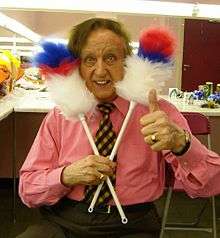 Colour photograph of Ken Dodd in a dressing room in 2007. He is holding two feather dusters against his head in a comical fashion.