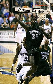 Kevin Garnett wearing black Timberwolves jersey as he jumps with the basketball towards the basket. Behind him are Washington Wizards players