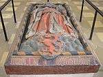 A stone tomb, with the effigy of a robed man on top, with a coat of arms at his feet displaying a red panther on a blue-and-gold striped background.