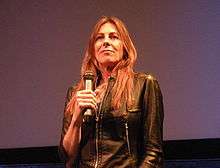 A brown-haired woman wears a black leather jacket with her hair flowing freely over her chest.