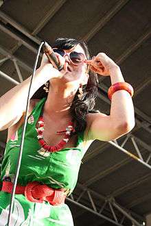 A black-haired girl in green dress with red belt singing on a microphone