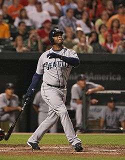 An dark skinned man wearing a gray baseball uniform with "SEATTLE" on the chest stands holding a baseball bat in his right hand as if having just taken a left-handed swing.