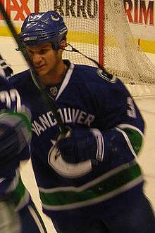 An ice hockey player in the midst of skating and holding his hockey stick upwards. His equipment is coloured blue and green.