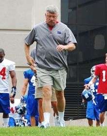 Photograph of Gilbride jogging onto a field with his head down wearing a grey New York Giants shirt and tan shorts
