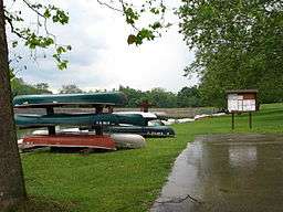 About a dozen canoes and small boats on a grassy shore of a lake at left, with a large puddle and a bulletin board and trees at right