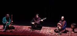 Kayhan Kalhor, Hossein Alizâdeh, and Majid Khalaj sit on a long Persian rug on a dimly-lit stage. The heads of Kalhor and Alizâdeh are lowered in concentration over their stringed instruments. Khalaj has closed eyes and a countenance of calm focus as his fingers strum the frame drum in his hand.