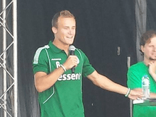 Fair-haired white man in a green shirt speaking into a microphone