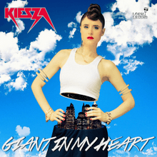A portrait of a young woman with tied-up red hair portrayed as mannequin split in half from her waist area. Her backdrop is a blue sky smitten with white clouds. She sport a white tank top, navy pants and bright red lipstick. Below her in large white font stands the title in capital letters 'Giant in My Heart', and in the top-left corner of the portrait stands the woman's name 'Kiesza' in small bold red font, also in capital letters.