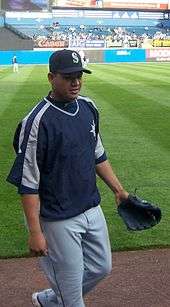 A young man wearing gray baseball pants, a navy-blue pullover windbreaker, and a black baseball glove (on his left hand) steps down from a dirt-and-grass field.