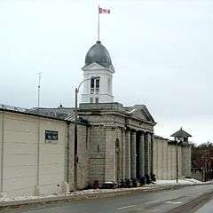 An ornate mock Greco-Roman entrance is viewed from the right by the viewer. The entrance is topped by a white stone tower, on top of which is a flagpole flying the Canadian flag, a red-white-red vertical tricolour with a red maple leaf in the middle. A tall, plain wall, lined on the far side with barbed wire, runs along the viewer's line of sight on either side of the entrance.