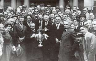 Black-and-white photo of a posed group of people, mostly adult males, mostly wearing overcoats, standing on the steps of a building with columns. A short man holding a silver cup stands at the front, next to a man wearing civic regalia.