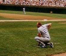 A man in a baseball uniform kneels looking in at the field.