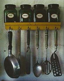 Various kitchen utensils.  At top: a spice rack with jars of mint, caraway, thyme, and sage.  Lower: hanging from hooks; a small pan, a meat fork, an icing spatula, a whole spoon, a slotted spoon, and a perforated spatula.
