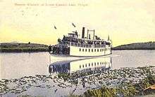 Colored-drawing postcard of a small steamboat packed with passengers, slowly moving through the lake's marshlands, with birds circling overhead