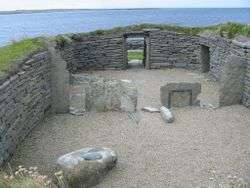 A small area of gravel is enclosed by a stone wall built into the surrounding grassy fields. Various large stones sit inside this enclosure and a low doorway has been constructed at the far end. Beyond the doorway there is a rocky foreshore and a body of water.