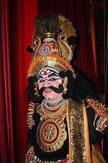 Person with painted eyes in yakshagana costume, as gold-spangled robe with red sheer scarf and spiked headress on gold crown