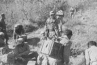 Unarmed men with boxes of supplies strapped to their backs at the bottom of a hill. In the background other men have begun the climb to the top.