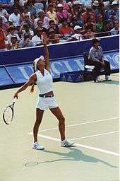 Anna Kournikova playing tennis in white outfit. Left hand is extended as if she has just tossed a ball and right hand is cocking back for the serve.
