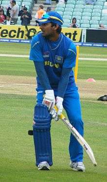 A side view of Kumar Sangakkara in blue clothes, holding a bat against the background of green grass and light yellow pitch.