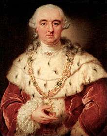 A white whigged man with a crimson coat and an ermine mantle holds a medallion in his hand. The medallion has a bright green stone in the center, and is encircled by a star-burst, and it hangs from a jewel-studded chain around his neck.