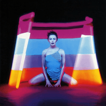 A body image of a woman (Kylie Minogue) inside of a cut cone with multi-colored lights. From top: Purple, Blue, mid-purple/blue, oranges and red. The woman is wearing a small blue mini dress with detail on the top left, whilst the floor has a reflection of the cone.