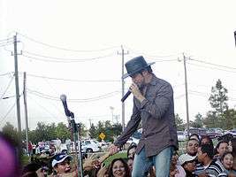 A man with a hat on his head, semi-inclined, wearing a colorful shirt and denim pants, holding a microphone in his left hand and holding the hand of a woman who hails from a crowd in the background. In the background you can see vehicles, poles and high voltage cables.